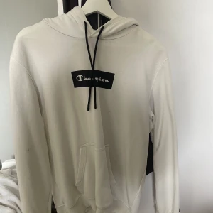 Vit hoodie från Champion - Vit hoodie från Champion med svart logga på bröstet och svarta snören i huvan. Klassisk känguruficka framtill och mjukt material. Perfekt för en avslappnad stil.