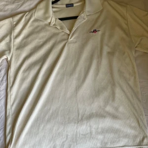 Vit pikétröja från Gant - Säljer en klassisk vit pikétröja från Gant med diskret randigt mönster och broderad logga på bröstet. Tröjan har krage och korta ärmar, perfekt för en clean och sportig stil.