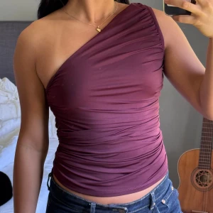 Vinröd one shoulder topp - Säljer en vinröd one shoulder topp med tight passform från Bikbok. Toppen har draperad effekt och är tillverkad i ett stretchigt material som sitter snyggt mot kroppen. Använd endast enstaka gånger. 100kr
