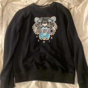 Svart sweatshirt med tigerbrodyr från Kenzo - Säljer en svart sweatshirt från Kenzo med ikonisk tigerbrodyr i blått, grått och vitt på bröstet. Tröjan har rund halsringning och långa ärmar. Perfekt för dig som vill sticka ut med en snygg detalj från ett känt märke.