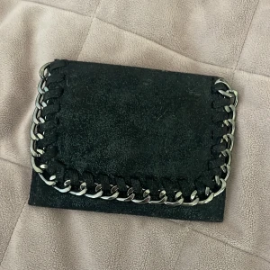 Svart plånbok med kedjedetalj - Säljer en svart plånbok i mocka med silverfärgad kedja runt kanten. Plånboken har magnetknäppning och flera fack för kort och sedlar. Perfekt accessoar för dig som gillar en tuffare stil. Säljer då den ej kommer till användning, vet ej märket på den💕 