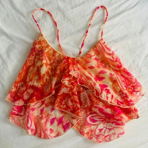 Färgglad volangig linnetopp från Bershka - Säljer en superfin och luftig linnetopp från Bershka i färgerna orange, rosa och vit. Toppen har tunna axelband och dubbla volanger, perfekt för varma dagar. Mönstret är livligt och ger en somrig känsla.