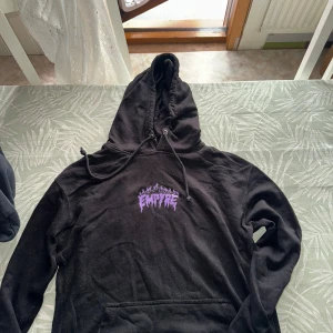 Svart hoodie från Empyre med lila tryck - Cool svart hoodie från Empyre med lila tryck framtill och stort motiv på ryggen. Tröjan har huva med dragsko och känguruficka. Perfekt för dig som gillar streetwear och vill sticka ut med ett unikt tryck.