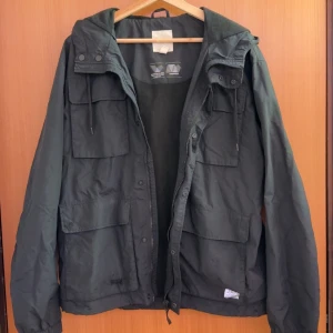 Mörkgrön vindjacka med huva från H&M - Säljer en mörkgrön vindjacka från H&M med huva och flera stora fickor framtill. Jackan har dragkedja och knappar, samt justerbar snörning vid huvan. Perfekt för blåsiga dagar och enkel att matcha med olika outfits.