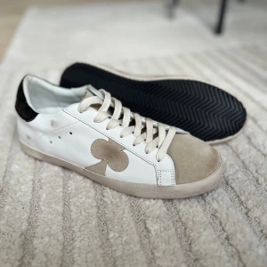 Vita sneakers från The Gallerist - Snygga vita sneakers från The Gallerist med beige mockadetaljer på tån och sidorna. Skorna har svarta detaljer på hälen med logga och klubbmönster längs baksidan. Klassisk snörning och platt sula. Perfekta för en clean och trendig look.