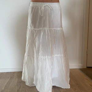 Vit långkjol med volanger och snörning - Superfin vit långkjol med volanger och snörning i midjan. Kjolen är luftig och har en avslappnad passform, perfekt för varma dagar. Tillverkad i ett lätt tyg som ger en skön känsla och snyggt fall. Lite genomskinlig på baksida.