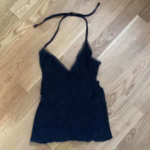 Svart spetsig korsett-topp med snörning - Supersnygg svart korsett-topp i spets med blommigt mönster och v-ringning. Toppen har smala axelband som knyts i nacken och snörning i ryggen för en justerbar passform. Perfekt för dig som vill sticka ut med en edgy och feminin look.