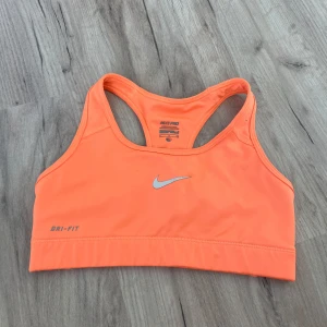 Orange sporttopp från Nike Pro - Sportig orange linnetopp från Nike Pro med klassisk racerback och silverfärgad logga framtill. Tillverkad i Dri-Fit-material som andas och håller dig torr under träningen. Perfekt för dig som vill ha en snygg och bekväm topp till gymmet eller löprundan.