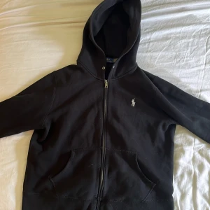Svart hoodie från Polo Ralph Lauren - Svart hoodie från Polo Ralph Lauren med dragkedja framtill och klassisk huva. Tröjan har långa ärmar, känguruficka och den ikoniska broderade loggan på bröstet. Perfekt för en chill och stilren look.