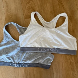 Calvin Klein sportiga linnetoppar - Två sportiga linnetoppar från Calvin Klein, en i ljusgrått och en i vitt. Båda har bred grå resår med logga och klassisk racerback. Perfekta för träning eller chill, i mjuk bomullsmix som sitter skönt mot kroppen.