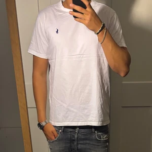 Vit t-shirt från Ralph Lauren - Säljer en klassisk vit t-shirt från Ralph Lauren med blå broderad logga på bröstet. T-shirten är i bomull, har rund hals och korta ärmar. Enkel och stilren modell som passar perfekt till jeans eller shorts.