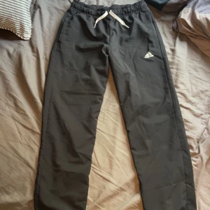 Svarta träningsbyxor från Adidas - Svarta träningsbyxor från Adidas med vit dragsko i midjan och klassisk Adidas-logga på låret. Byxorna har elastisk midja och är tillverkade i ett lätt och bekvämt syntetmaterial, perfekta för sport eller chill. Snygg och enkel design som funkar till det mesta.