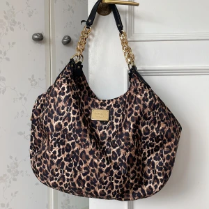Leopardmönstrad axelväska Victoria's Secret - Stor och rymlig axelväska från Victoria's Secret med leopardmönster i brunt och svart. Väskan har guldfärgade kedjedetaljer på axelremmen och en tydlig logoplatta framtill. Tillverkad i syntetmaterial och stängs med dragkedja.