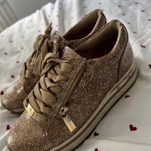 Glittriga guld sneakers med dragkedja - Snygga sneakers i beige med glittrigt rutmönster och guldiga detaljer. Skorna har breda snören, dragkedja på sidan och platt sula. Perfekta för dig som vill sticka ut med en unik och trendig look.