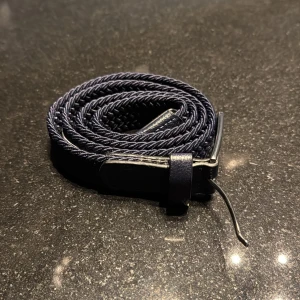 Mörkblått flätat bälte från Kappahl - Snyggt mörkblått bälte från Kappahl med flätad design och detaljer i skinn. Bältet har en klassisk silverfärgad metallspänne och är smalt, vilket gör det lätt att styla till olika outfits. Perfekt för dig som gillar stilrena accessoarer.