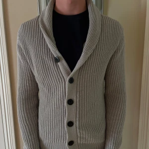 Beige stickad kofta med sjalkrage - Snygg beige stickad kofta med grov ribbad struktur och sjalkrage. Koftan har svarta knappar framtill och lång ärm, perfekt för lager-på-lager. Passar dig som gillar en klassisk men chill stil.