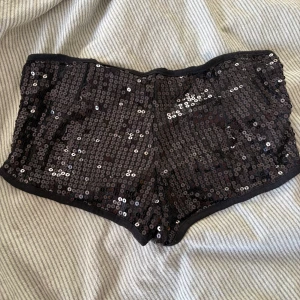 Svarta paljettshorts/hotpants S/M - Glittriga svarta hotpants täckta med paljetter, perfekta för dig som vill sticka ut. De är stretchiga och mjuka tack vare viskos och elastan, med en tight och kort passform. Snygga att matcha med en enkel topp för en cool look.