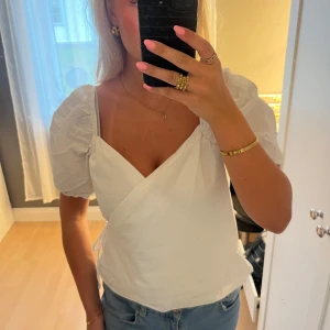 Vit omlottopp med puffärm - Superfin vit omlottopp med puffiga korta ärmar och knyt i sidan. Toppens v-ringning och voluminösa axlar ger en trendig och feminin look. Perfekt att styla med jeans eller kjol för en fräsch vibe.