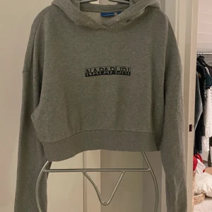   Hoodie från Napapijri! - Snygg grå croppad hoodie från Napapijri med broderad logga på bröstet. Tröjan har huva, långa ärmar och ribbade muddar. Perfekt för en chill och avslappnad stil. Tillverkad i mjuk bomullsmix som känns skön mot huden. Är endast använd och tvättad vid 2 tillfällen. Så är i nyskick. Snören saknas.                 Säg till om du vill ha flera bilder! 🌸