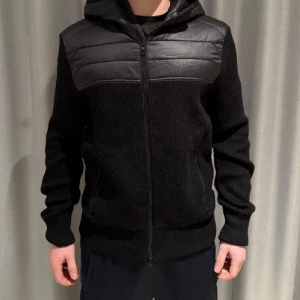 Svart stickad jacka med huva - Svart jacka med stickade ärmar och kropp, samt quiltade detaljer i syntetmaterial över axlar och bröst. Jackan har huva, dragkedja framtill och två fickor med dragkedja. Perfekt för dig som gillar en sportig och stilren look.