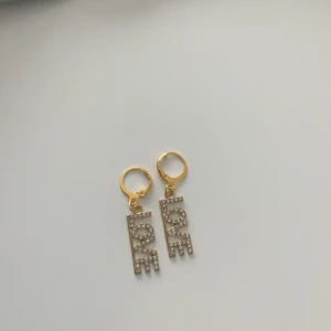 Love örhängen  - Säljer ett par superfina örhängen i guldton med gnistrande strass-stenar. Hängena bildar ordet “LOVE” med en modern och unik design, perfekt för att addera lite glamour till din outfit.