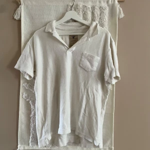 Vit frotté pikétröja från OAS - Vit pikétröja från OAS i 100% bomullsfrotté, perfekt för sommaren. Tröjan har korta ärmar, öppen krage och en bröstficka med broderad palm. Skön och avslappnad passform, riktigt chill vibe för strand eller city.