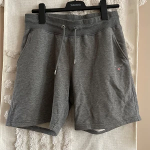 Grå mjukisshorts från GANT - Grå mjukisshorts från GANT med broderad logga på vänster sida. Shortsen har snörning i midjan, två sidofickor och en bakficka. Sköna och avslappnade med normal passform, perfekta för chill dagar eller träning.