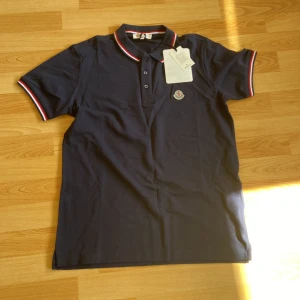 Mörkblå Moncler pikétröja - Snygg mörkblå pikétröja från Moncler med klassisk krage och knappar framtill. Röd, vit och blå rand på krage och ärmslut samt broderad Moncler-logga på bröstet. Tillverkad i bomull för en skön och stilren känsla.