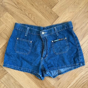 Blå jeansshorts  - Klassiska blå jeansshorts med hög midja, raka ben och två fickor fram. Snygga detaljer med bälteshällor och knappstängning. Perfekta för sommaren och lätta att matcha med allt. Materialet är mjuk denim i bomull.