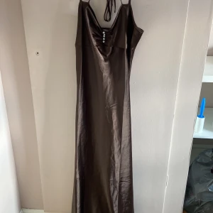 Brun långklänning från LingaDore - Snygg brun långklänning från LingaDore i glansigt satinliknande material. Klänningen har smala axelband, djup urringning med dekorativa stenar framtill och en elegant, figurnära passform. Perfekt för dig som vill sticka ut med en stilren look.