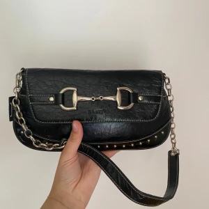 Svart axelväska från Biaggio med kedja - Cool svart axelväska från Biaggio i skinnimitation med silverfärgade metalldetaljer och kedjerem. Väskan har ett hästinspirerat spänne framtill och nitar längs kanten. Perfekt storlek för det viktigaste och ger en edgy vibe till din outfit.❤️🥳 Använt men fint skick, se bilder.