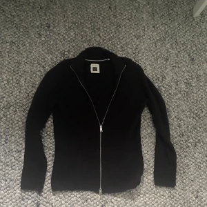  ribbad kofta från Marc O'Polo - Grå ribbstickad kofta från Marc O'Polo med hel dragkedja framtill och hög krage. Tillverkad i ett mjukt, organiskt material som känns skönt mot huden. Perfekt för lager-på-lager och enkel att matcha med olika outfits.