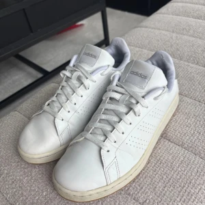 Vita Adidas sneakers  - Klassiska vita Adidas sneakers i skinn med diskreta perforerade ränder på sidorna och ljusgrå detalj vid hälen. Skorna har snörning och en platt sula, perfekt för en clean och stilren look. Adidas-logga syns på tunga och häl.