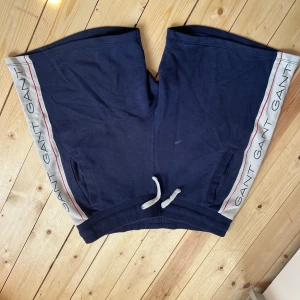 Marinblå shorts från Gant med snörning - Snygga marinblå shorts från Gant med vita och röda detaljer längs sidorna där det står GANT. Shortsen har snörning i midjan och två fickor framtill. Perfekta för sommaren och chill dagar.