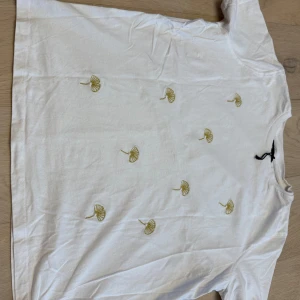 Vit t-shirt med broderade detaljer - Vit t-shirt i bomull med korta ärmar och små broderade blommor i gult och pärldetaljer över hela framsidan. Enkel och fräsch design som passar perfekt till jeans eller kjol. Skön och luftig passform.