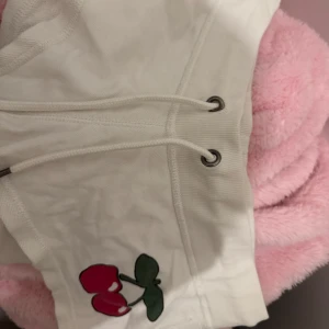 Vita shorts med körsbär - Supersöta vita shorts med snörning framtill och ett broderat körsbärsmotiv på ena benet. Mjukt bomullsmaterial och bekväm passform, perfekta för sommardagar. Liten etikett med Peachy-logga bak. Passar både som pyjamas shorts eller en sommardag ute. Dom är korta. Köptes i Spanien☀️🍒