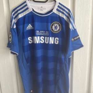 Chelsea 2012 Drogba matchtröja Adidas - Chelsea FC hemmatröja från Champions League-finalen 2012 med Drogba 11 på ryggen. Blå med vita detaljer, Adidas-logga, klubbmärke och Samsung som sponsor. Kortärmad med randigt mönster i olika blå nyanser och vita axelband. Tillverkad i lätt funktionsmaterial.