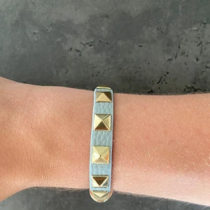 Valentino liknande armband  - Snyggt ljusblått armband i skinn med stora fyrkantiga guldnitar längs hela bandet. Justerbar längd med flera hål och guldfärgat spänne. Perfekt accessoar för att ge din outfit en edgy touch.