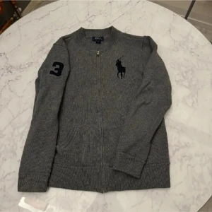 Grå stickad kofta från Polo Ralph Lauren - Snygg grå stickad kofta från Polo Ralph Lauren med dragkedja framtill, ribbade muddar och fickor. Stor svart logga på bröstet och siffra 3 på ärmen ger en sportig vibe. Perfekt för lager-på-lager under höst och vinter.