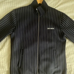 Randig bomberjacka från Les Deux - Snygg svart bomberjacka från Les Deux med vita tunna ränder. Jackan har hög krage, dragkedja framtill och två fickor. Stilsäker och modern look med diskret logga på bröstet. Perfekt för dig som gillar streetstyle och vill sticka ut.