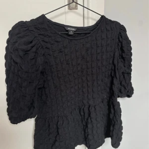 Svart strukturerad blus från Monki - Supersnygg svart blus från Monki i storlek XS med puffiga ärmar och coolt quiltad struktur. Modellen har en lätt volang nertill och trekvartsärm. Perfekt för dig som gillar unika detaljer och vill sticka ut lite extra.