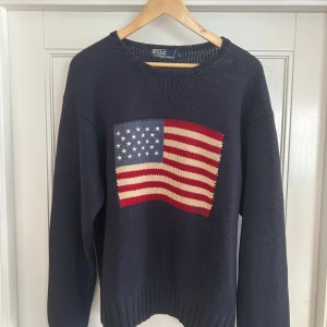 Mörkblå stickad tröja Polo Ralph Lauren vintage från 90 talet - Säljer en mörkblå stickad tröja från Polo Ralph Lauren med en stor amerikansk flagga framtill. Tröjan har rund halsringning, ribbade muddar och lång ärm. Perfekt för dig som vill sticka ut med en klassisk och ikonisk look. köpt i början av 90-talet. 🧶 Material: 100 % bomull 📏 Storlek: L (passar även M för en lite oversize look) 📦 Skick: Nästan oanvänd – inga hål, fläckar eller noppning.En riktig vintagepärla för samlare eller dig som älskar klassisk Ralph Lauren-stil. 