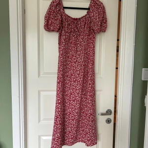 Röd blommig långklänning med puffärm - Köpt second hand för 150 kr. Superfin röd långklänning med vita blommor, puffiga kortärmar och söt knytning framtill. Materialet är mjukt och lätt. Har en slits på ena benet (bild 4). Midiklänning. 