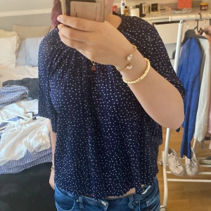 Mörkblå prickig blus från Indiska - Supersöt mörkblå blus från Indiska med vita prickar och lös passform. Blusen har korta ärmar och rynkade detaljer vid halsen som ger en chill och luftig vibe. Perfekt att styla med jeans eller kjol för en fräsch look.