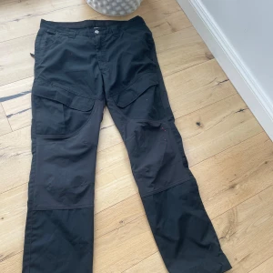 Svarta cargopants från Kansas - Svarta cargopants från Kansas med stora fickor på sidorna och förstärkta knän. Byxorna är tillverkade i slitstark polyesterblandning och har klassisk rak passform. Perfekta för dig som gillar funktionella och stilrena arbetsbyxor.