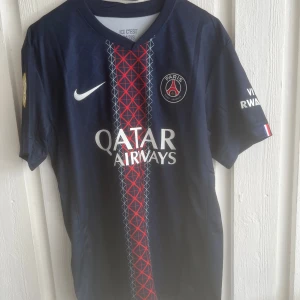 PSG Dembele matchtröja Nike blå - Paris Saint-Germain fotbollströja med O. Dembélé och nummer 10 på ryggen. Djupblå med röd och vit grafisk detalj i mitten, PSG-emblem på bröstet och Qatar Airways-tryck. Korta ärmar, officiella FIFA-märken på ärmen. Sval och lätt polyester.