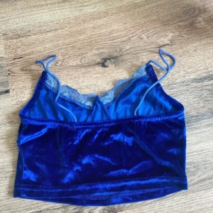 Blå sammetslinne med spetsdetalj - Supersnygg croppad linne-topp i djupblå sammet med tunna axelband och spetskant vid urringningen. Perfekt för dig som vill sticka ut med en lyxig känsla och cool vibe. Sitter tajt och har en mjuk, glansig finish som fångar ljuset.