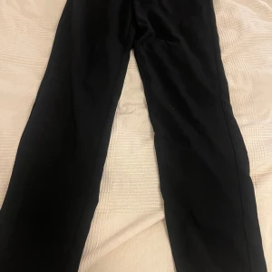 Svarta kostymbyxor från Gina Tricot - Klassiska svarta kostymbyxor från Gina Tricot med raka ben och midjehällor. Byxorna har dragkedja och knapp framtill samt diskreta fickor. Perfekta för en clean och stilren look.