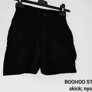 Svarta cargo shorts från Boohoo - Svarta cargo shorts från Boohoo med hög midja och stora fickor på sidorna. Klassisk knapp och dragkedja framtill, bälteshällor och en loose fit. Perfekta för en chill och trendig look under sommaren.  Nyskick så ingenting att anmärka på