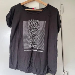 Joy Division svart t-shirt - Svart t-shirt med ikoniskt vitt tryck från Joy Divisions album Unknown Pleasures. Klassisk rund hals och korta ärmar. Perfekt för dig som gillar bandmerch och vill ha en stilren, tidlös look.